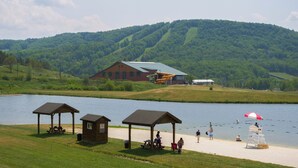 A lakeside setting with picnic shelters, a lifeguard stand, and a building with a green roof.