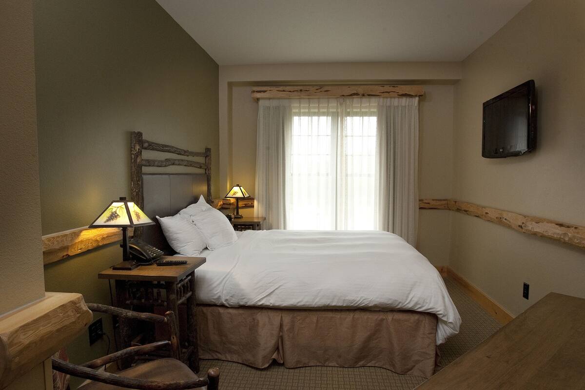 A hotel room with a large bed, two bedside lamps, a wooden headboard, a television mounted on the wall, and a chair.