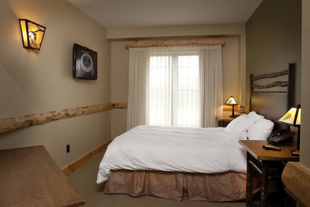 A hotel room with a large bed, a wooden headboard, a bedside table with a lamp, and a wall-mounted telephone.