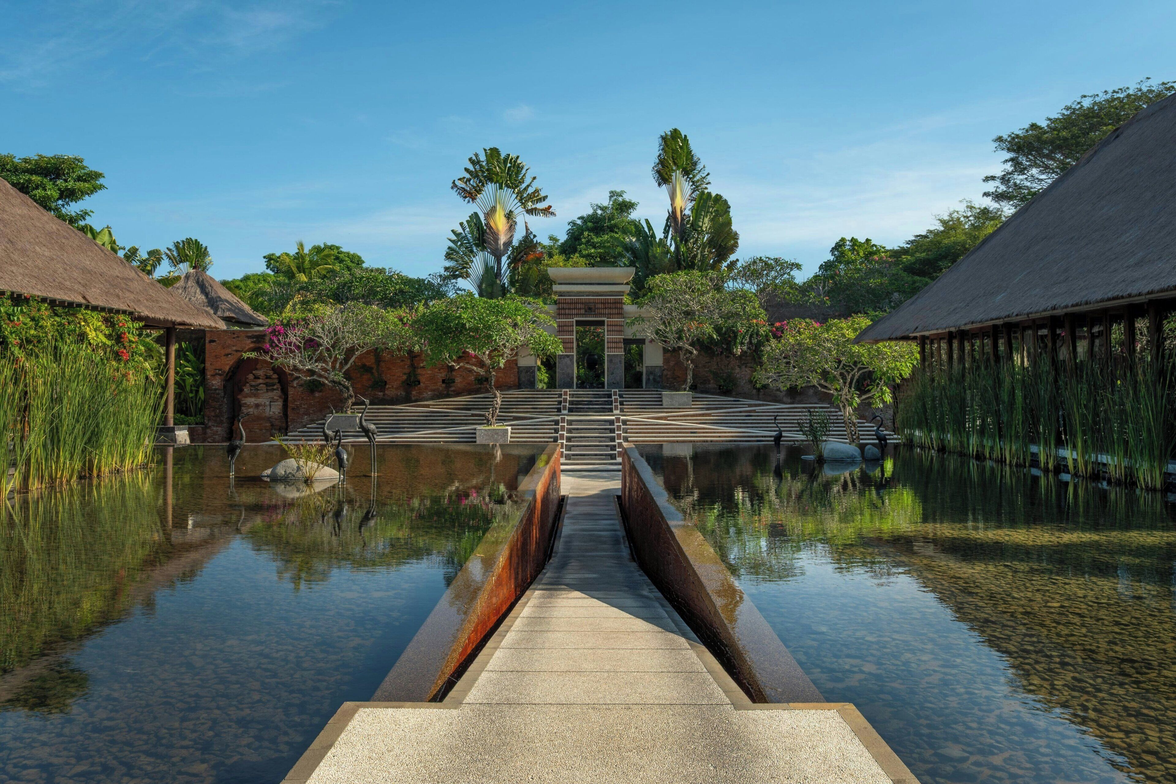 A tranquil waterway leading to a traditional building with a thatched roof, surrounded by lush greenery and a clear sky.