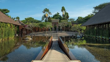 A tranquil waterway leading to a traditional building with a thatched roof, surrounded by lush greenery and a clear sky.