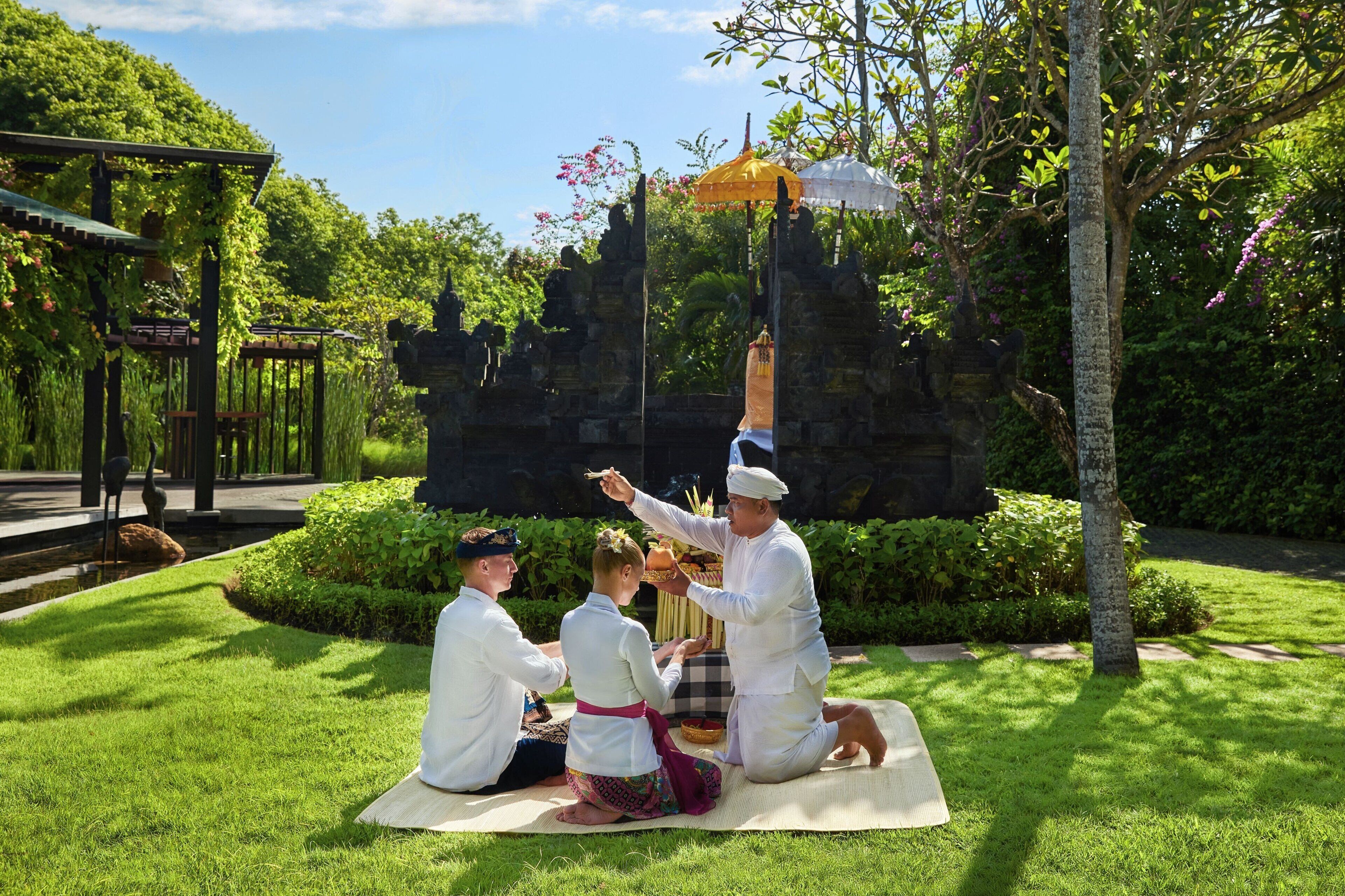 A group of people in traditional attire sitting on a mat outdoors, with a temple structure in the background.