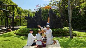 A group of people in traditional attire sitting on a mat outdoors, with a temple structure in the background.