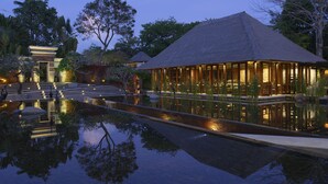 Un resort con una piscina reflectante, una estructura de techo de paja y un edificio tradicional con escalones que lo conducen.