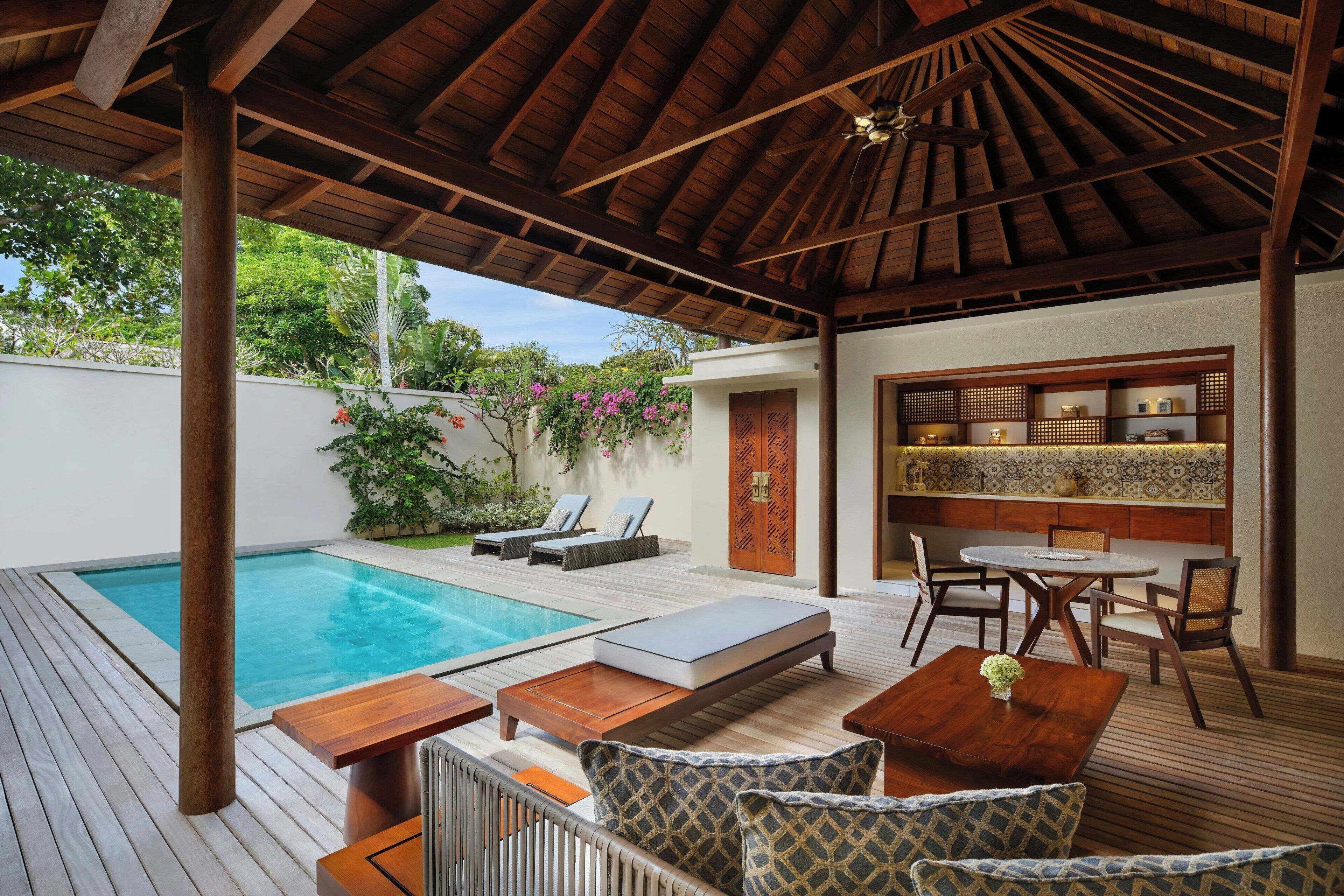 Uma área externa coberta com piscina, espreguiçadeiras, mesa de madeira e um espaço de estar com cadeiras e uma pequena mesa.