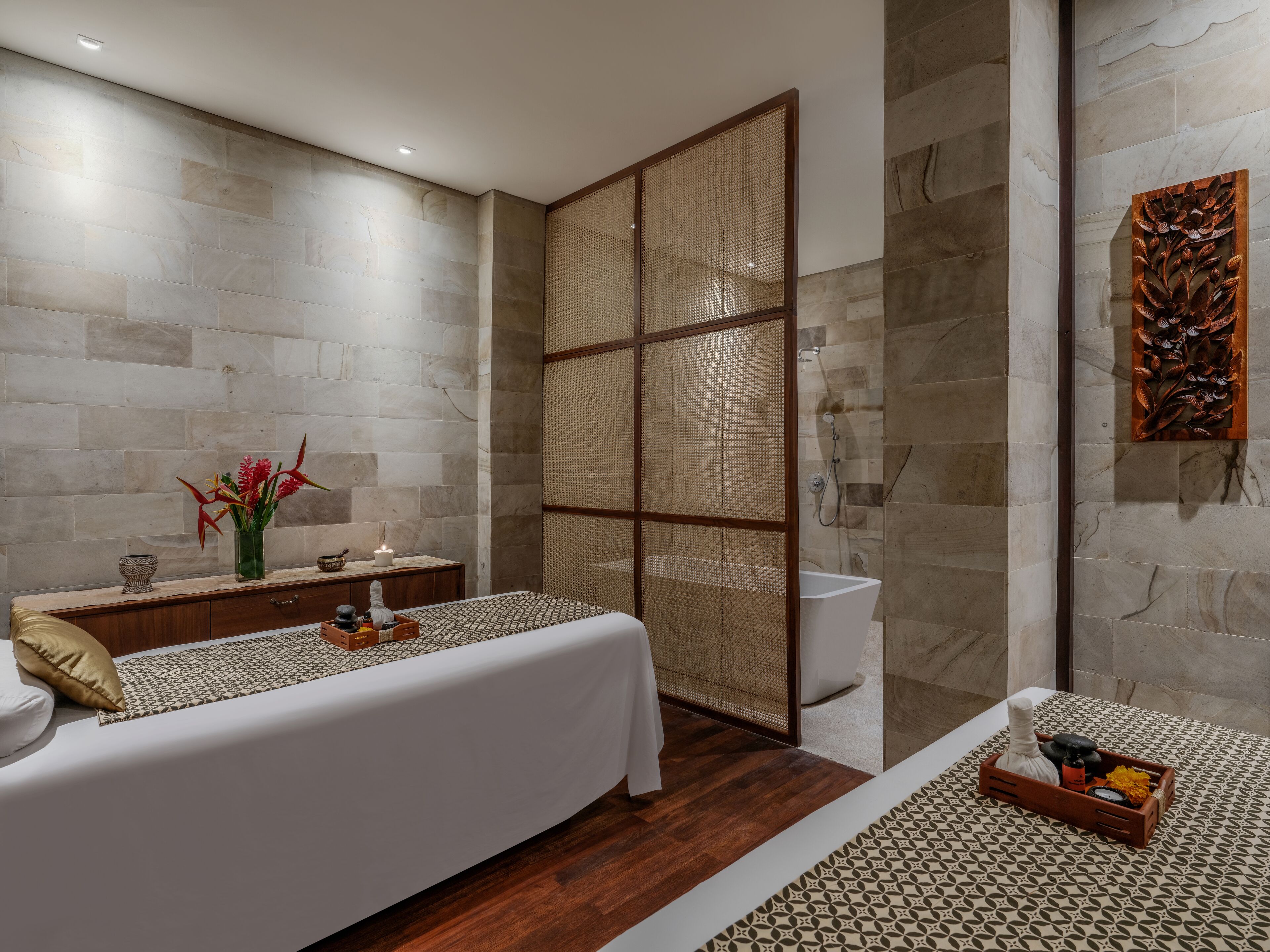 A spa room with a massage table, a small bedside table with a candle, a decorative wall piece, and a shower area with a stone wall.