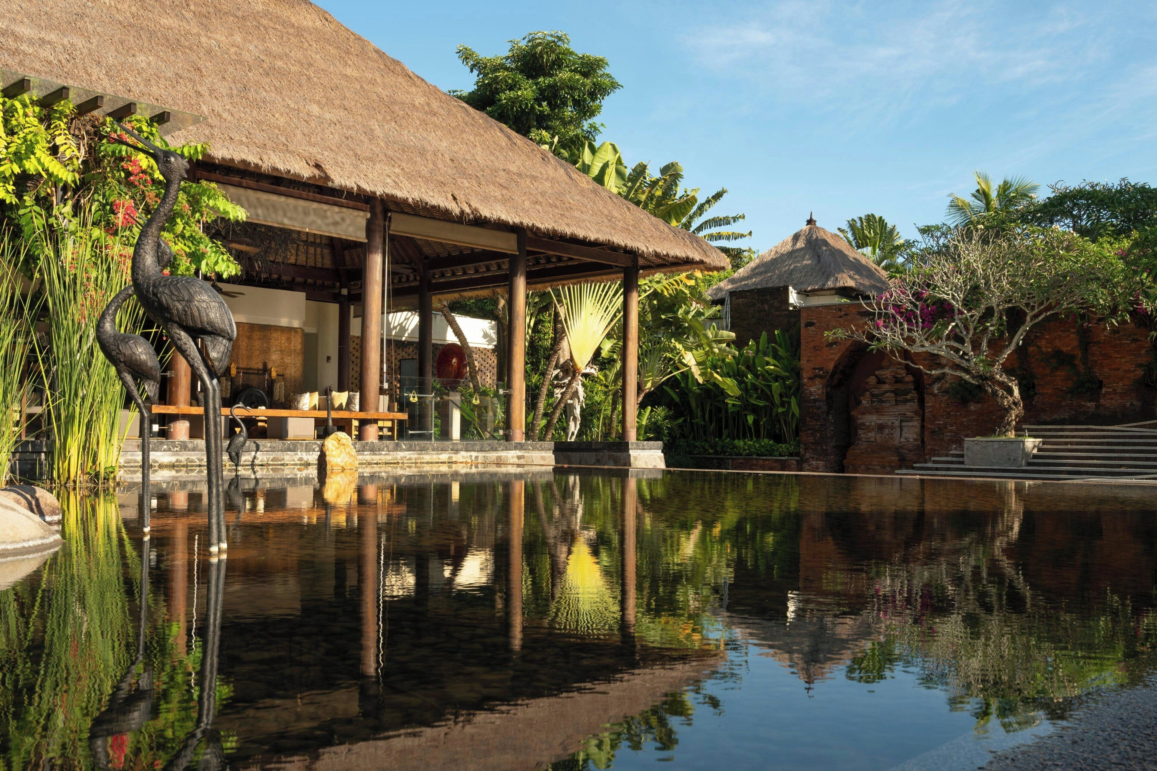 A tranquil pool area with a thatched-roof structure, a stone bridge, and lush tropical vegetation.