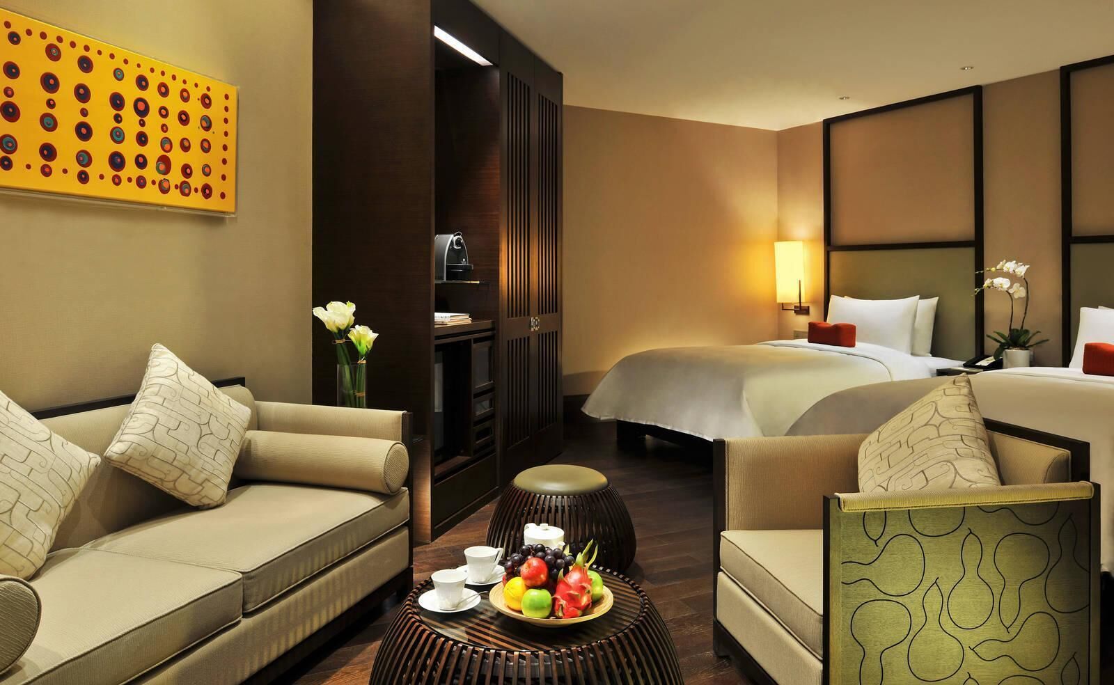 A hotel room with a sofa, a chair, a small table with fruit, a bed, and a nightstand.