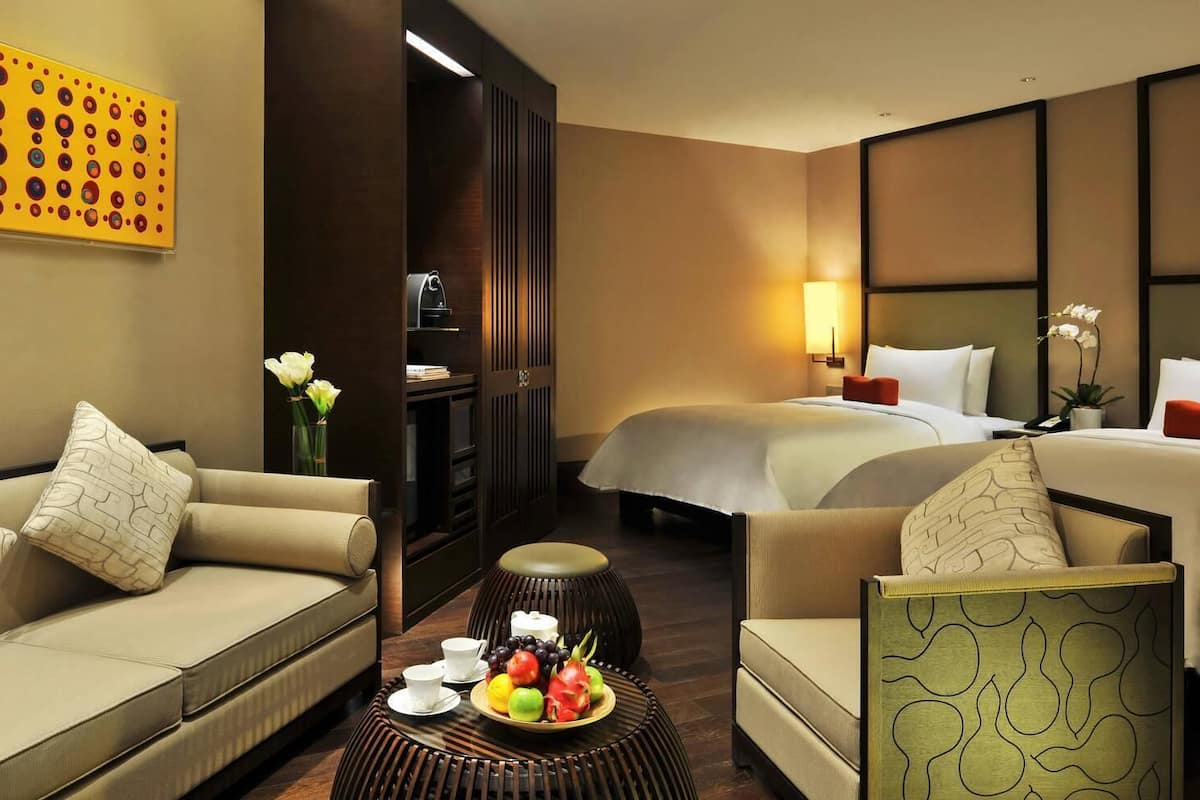 A hotel room with a sofa, a chair, a small table with fruit, a bed, and a nightstand.