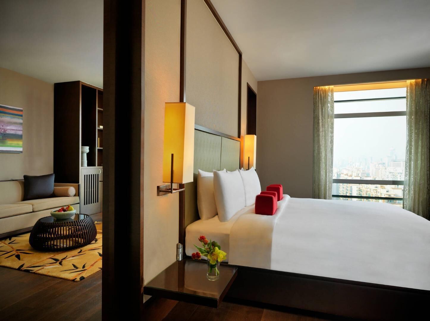 A hotel room with a large bed, a sofa, a small table with a vase, and a window with a city view.