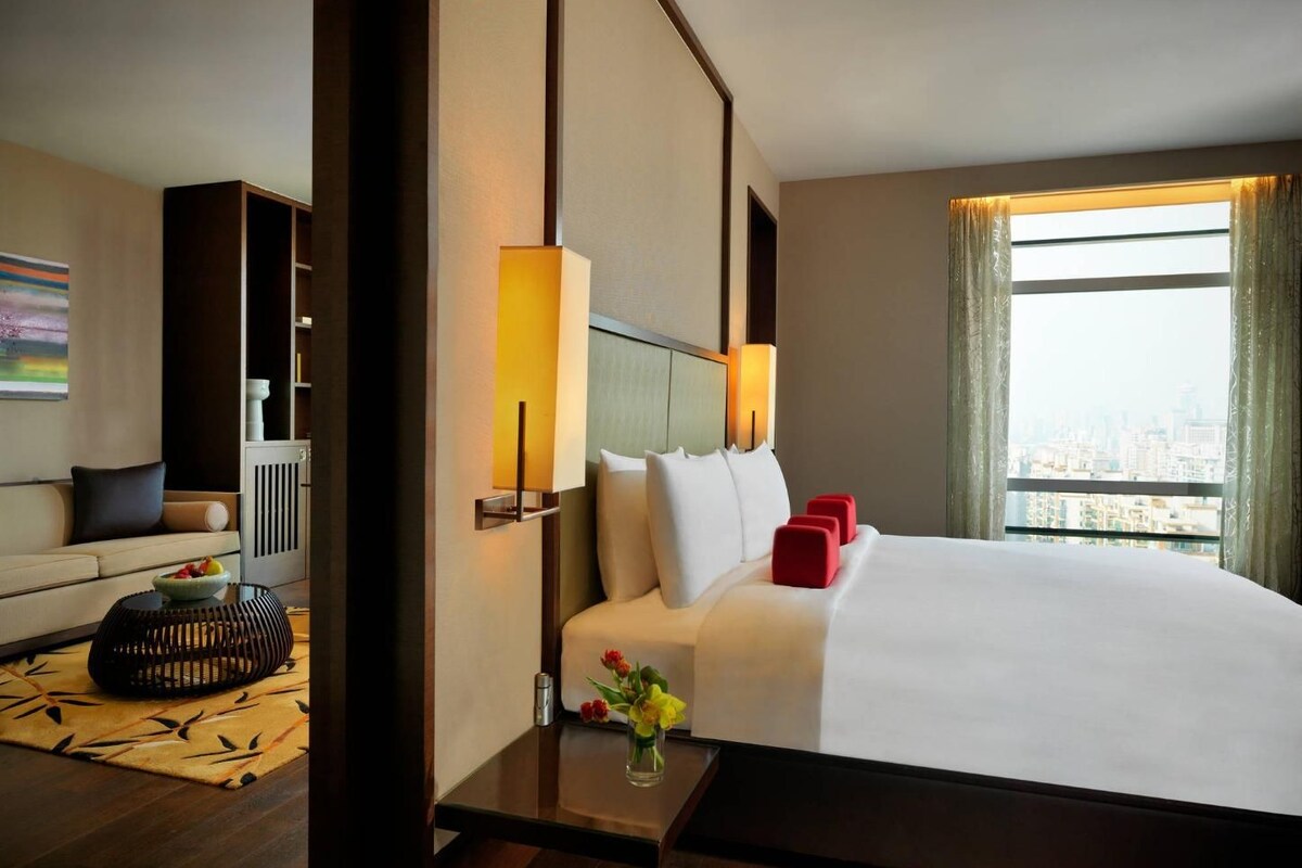 A hotel room with a large bed, a sofa, a small table with a vase, and a window with a city view.