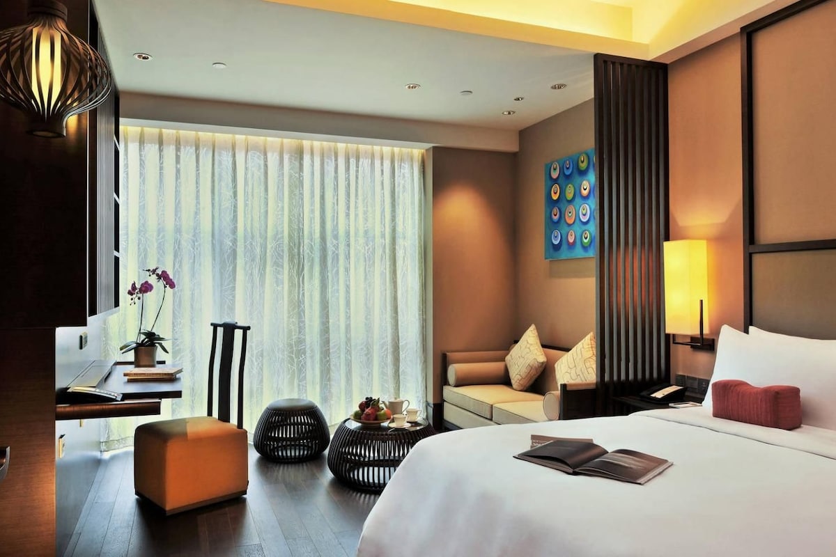 A hotel room with a bed, a desk with a computer, a sofa, a TV, and a window with curtains.