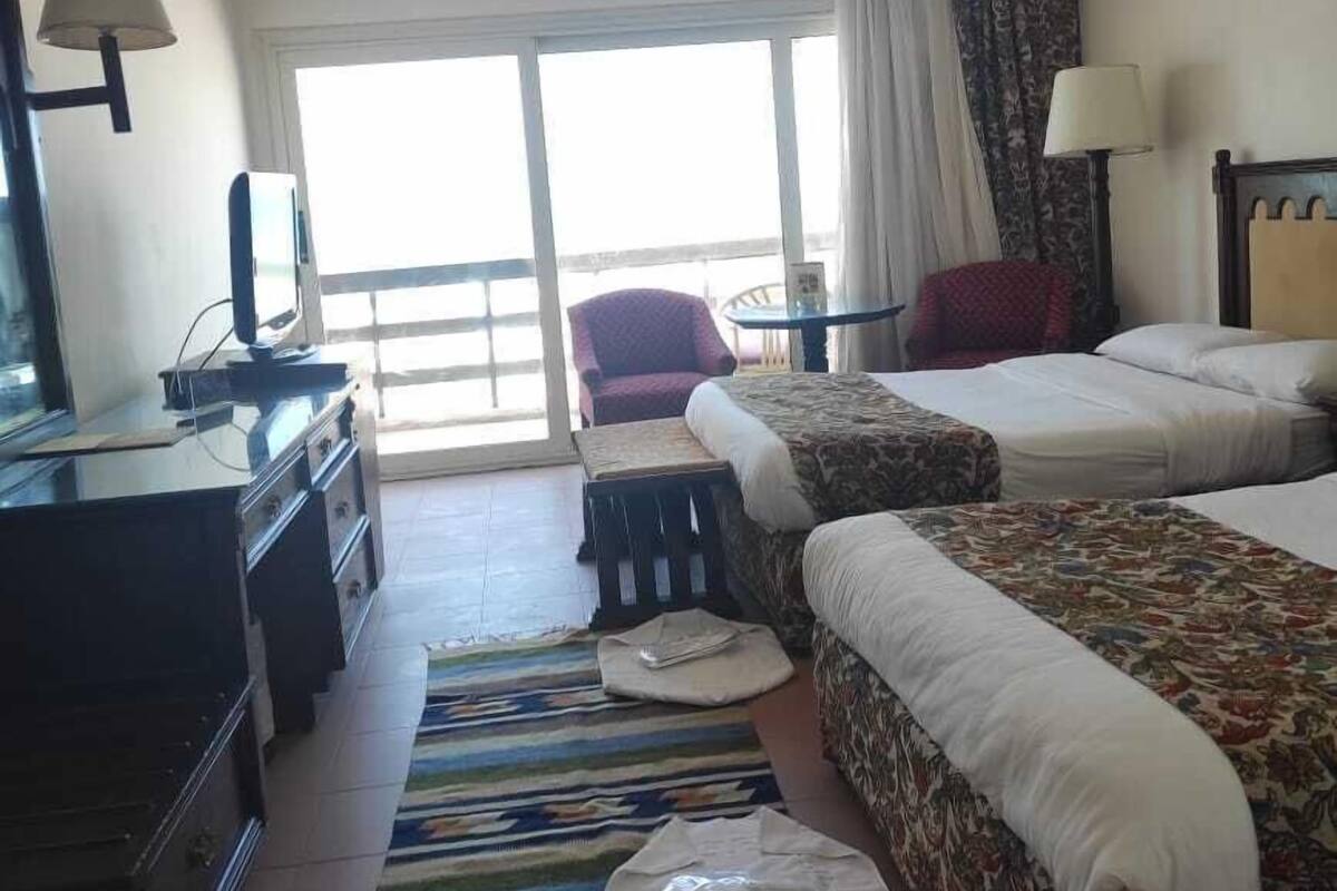 Quarto de hotel com duas camas, uma escrivaninha, uma cadeira e uma janela grande.