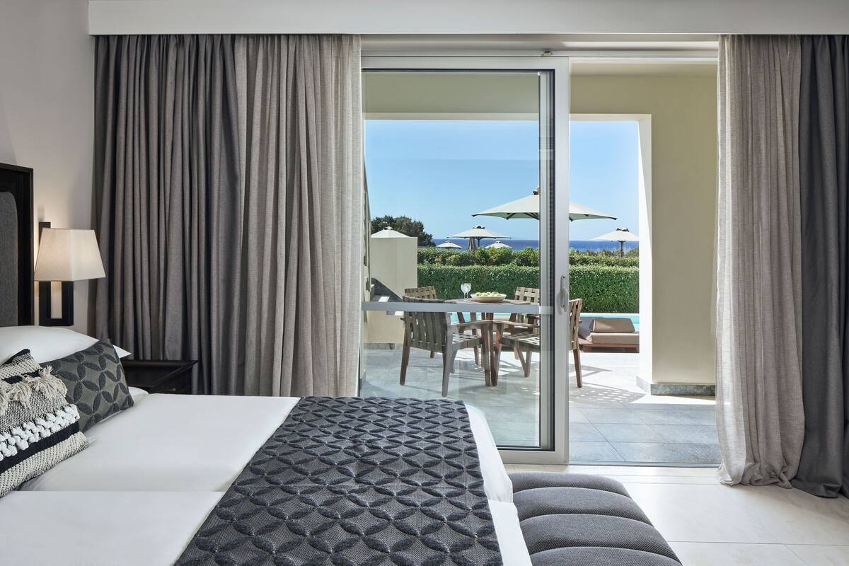 Ein Hotelzimmer mit einem Bett, einem Nachttisch, einer Lampe und Blick auf eine Außenpatio mit Sonnenschirmen und einem Pool.