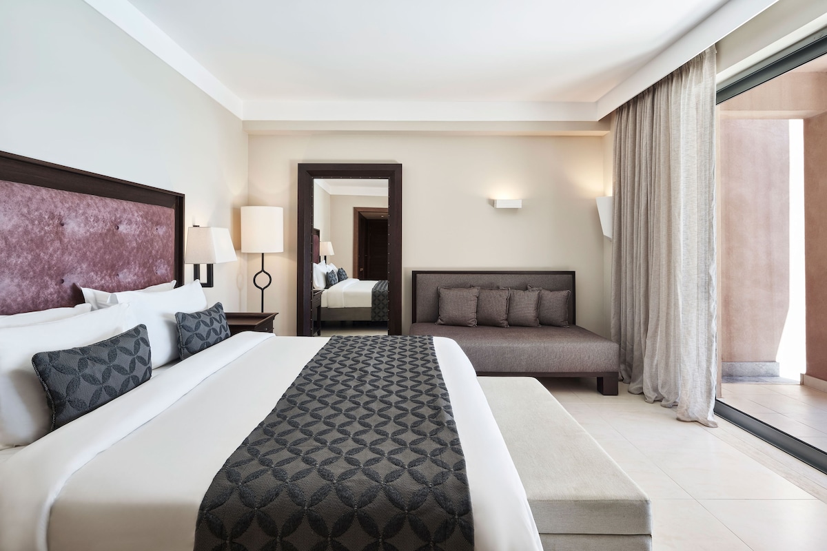 A hotel room with a large bed, a smaller sofa, a nightstand with lamps, and a view of the outside.