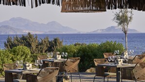 Een terras met rieten stoelen en tafels, met op de achtergrond bergen en de zee.