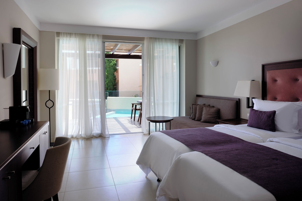 A hotel room with a bed, bedside table, lamp, and a view of a pool area.