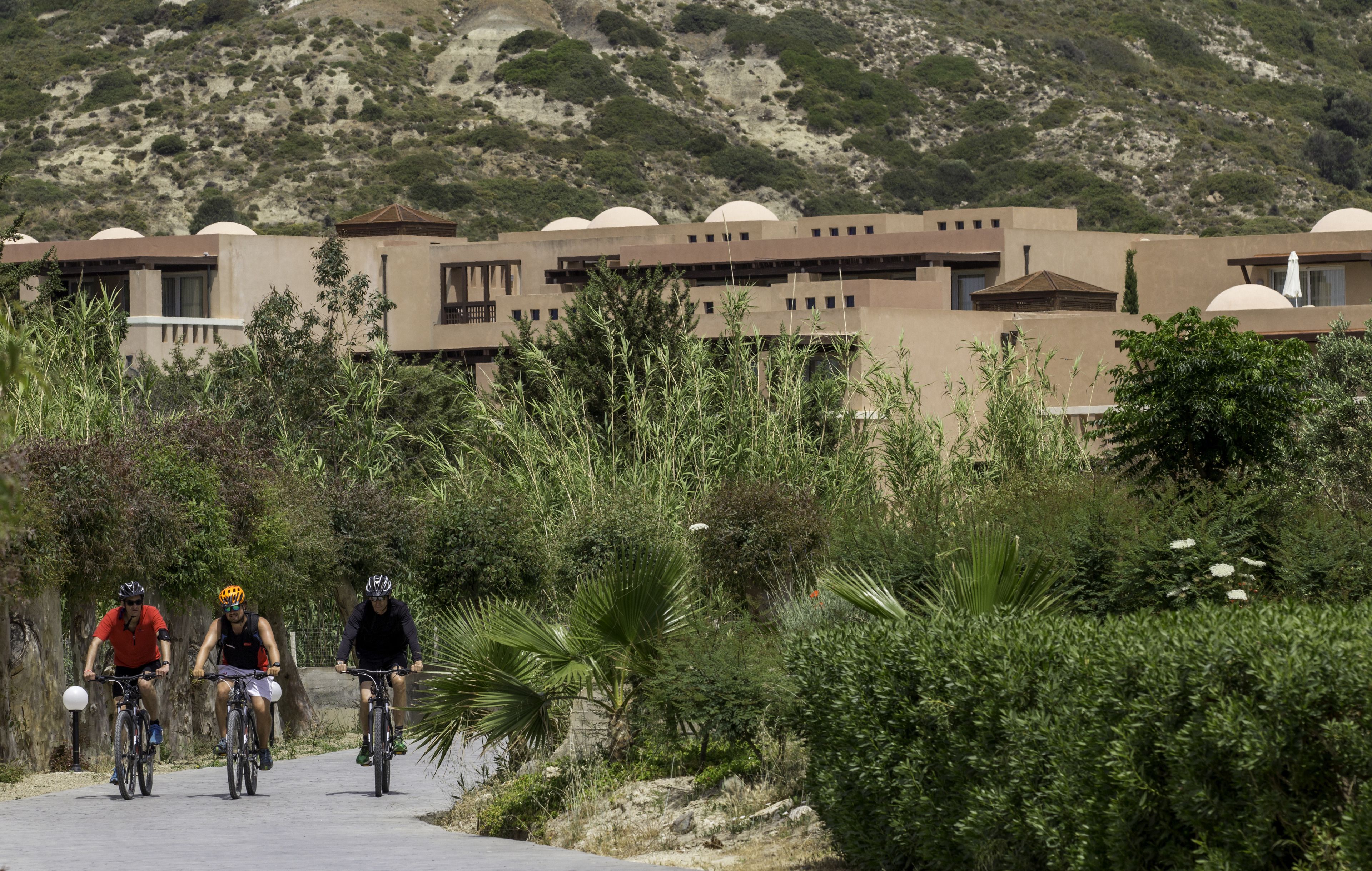 A pathway with three cyclists, surrounded by lush greenery and a building with multiple dome-shaped structures in the background.