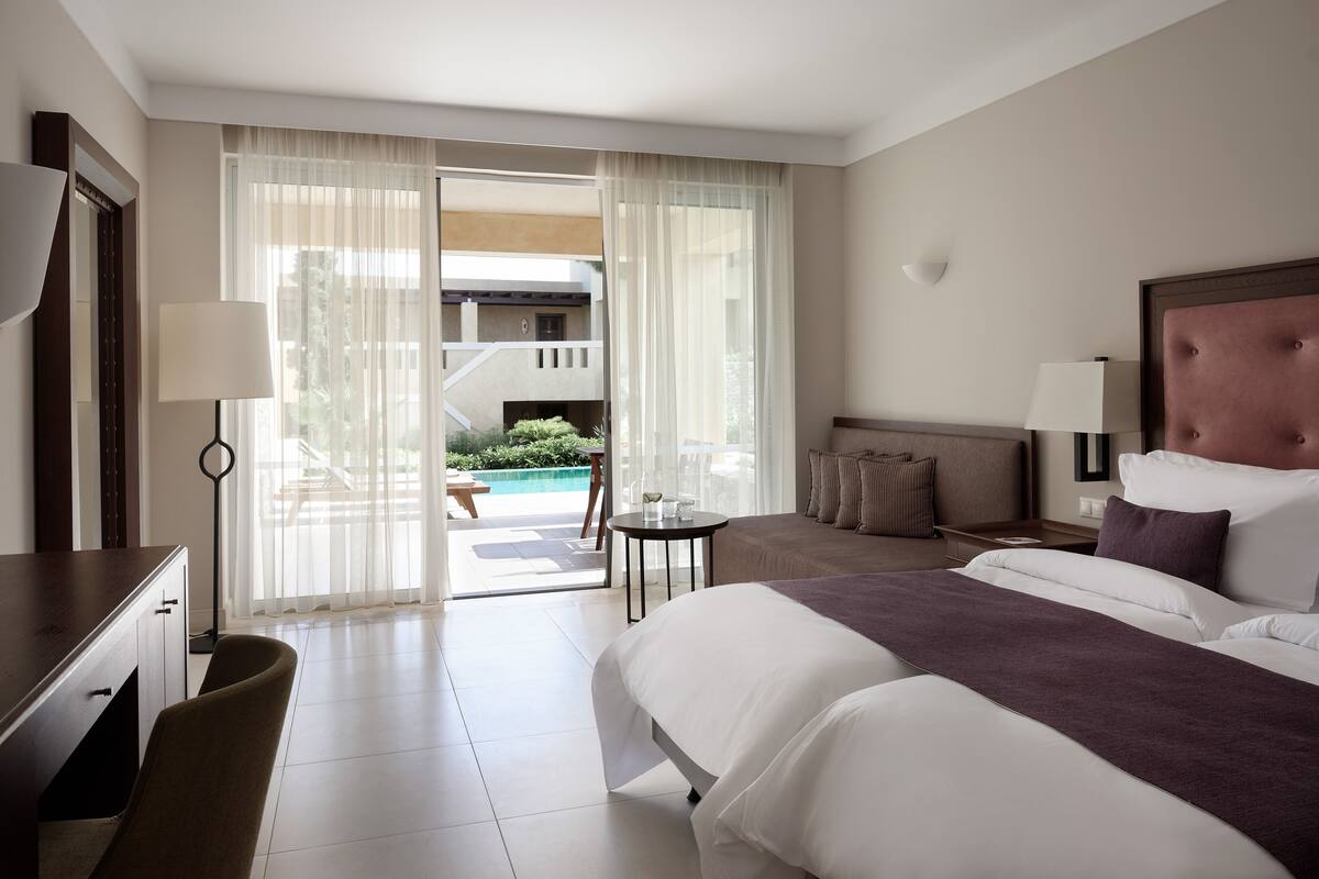 Ein Hotelzimmer mit Bett, Schreibtisch, Stuhl, Nachttisch, Lampe und Blick auf den Poolbereich.