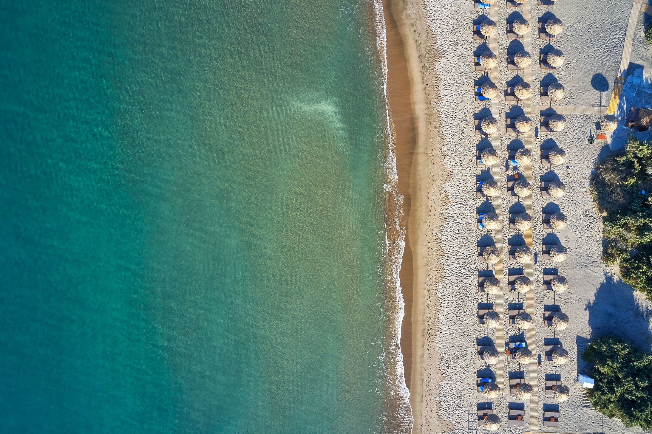 Eine Vogelperspektive zeigt einen Strand mit Reihen von Sonnenschirmen und Liegestühlen, was auf ein Resort hindeutet.