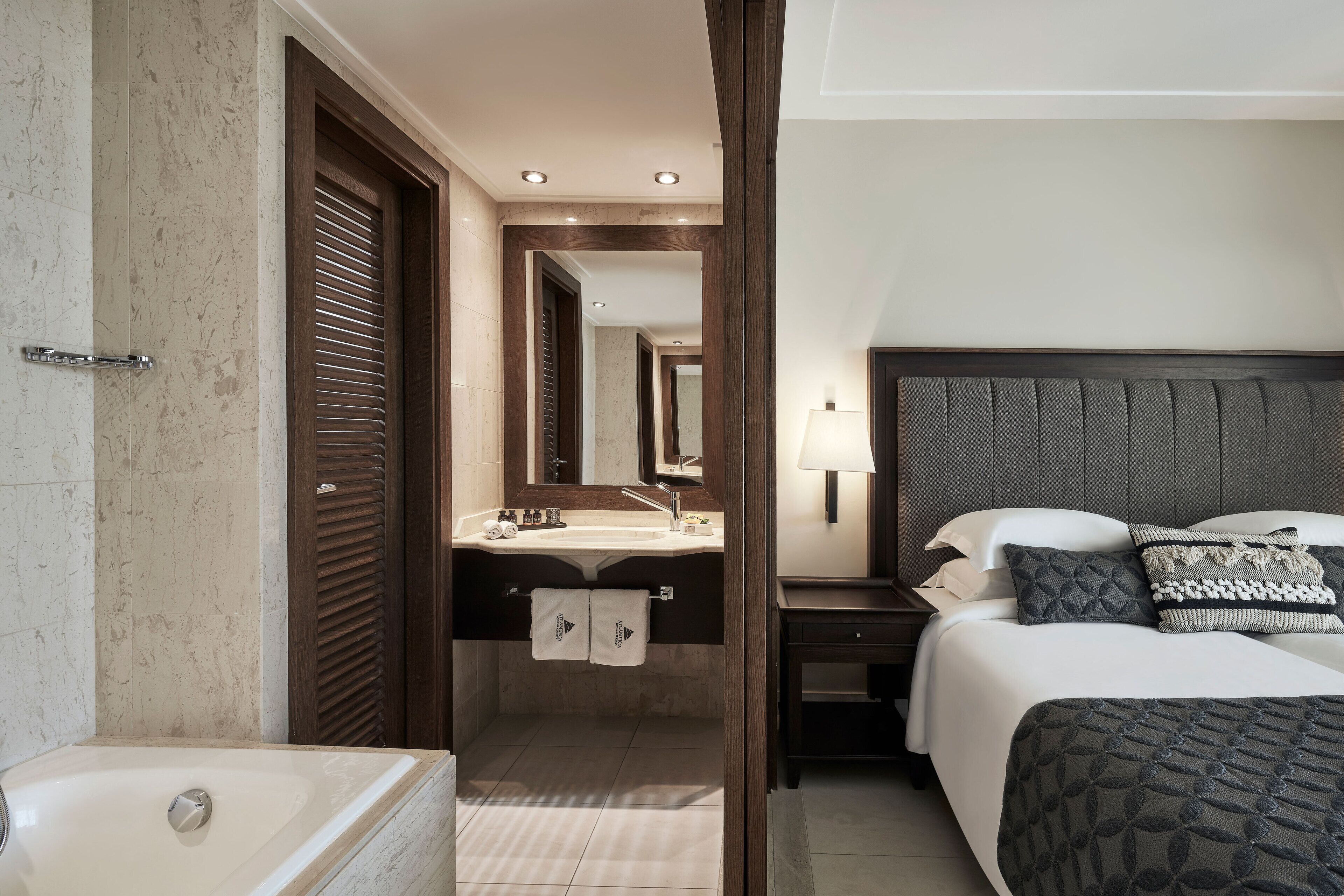A hotel room with a large bed, a bedside table, a nightstand, a bathroom with a bathtub, and a mirror.