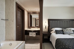 A hotel room with a large bed, a bedside table, a nightstand, a bathroom with a bathtub, and a mirror.