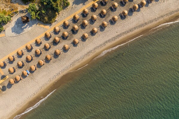 Vue aérienne d’une plage bordée de rangées de parasols en chaume et d’une promenade de sable.