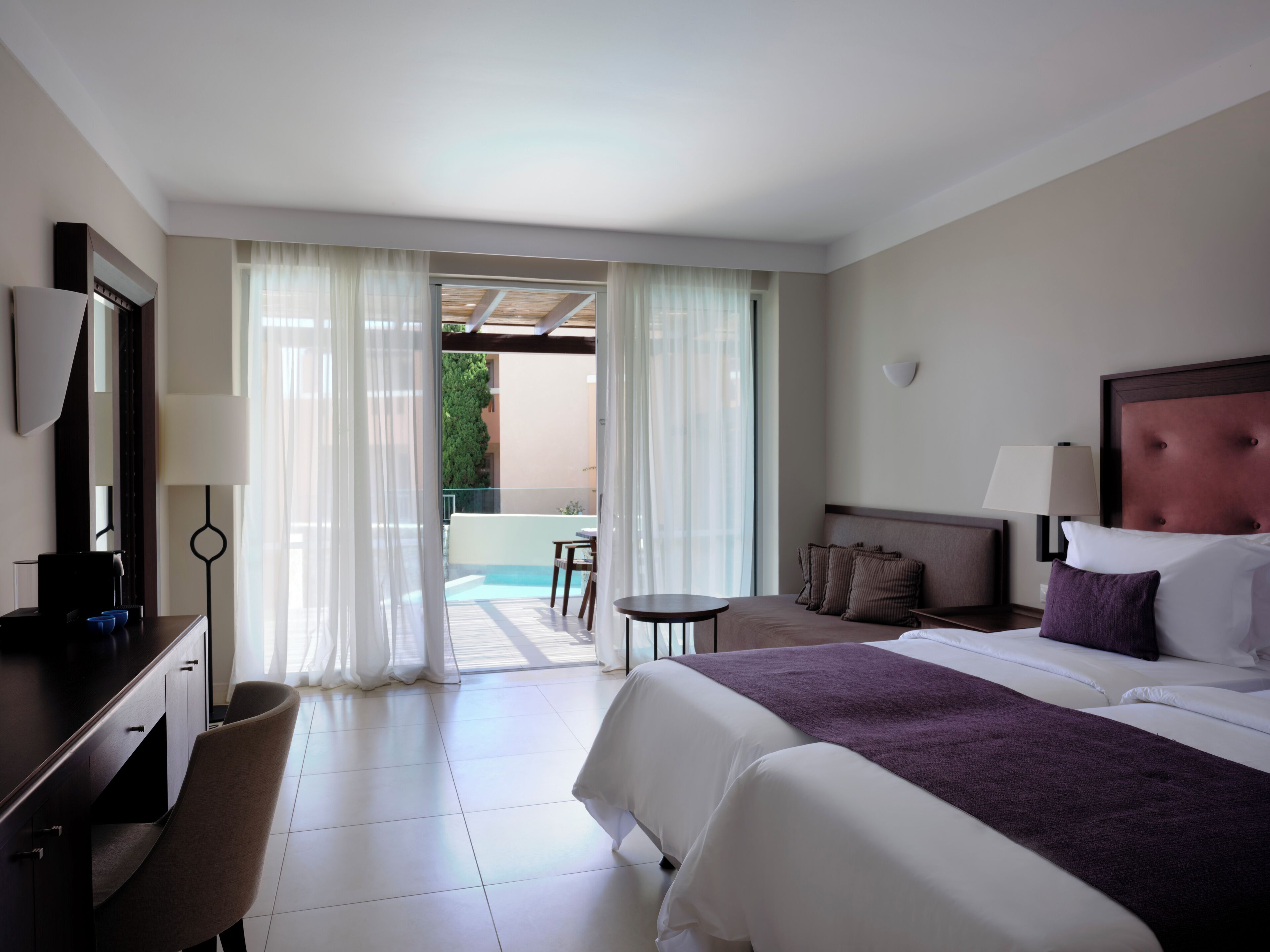 Ein Hotelzimmer mit Bett, Schreibtisch, Stuhl, einem kleinen Tisch und Blick auf einen Poolbereich im Freien.