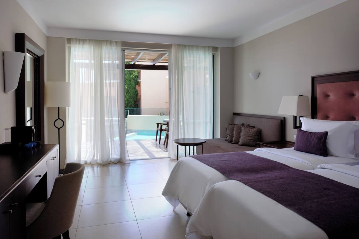 Ein Hotelzimmer mit Bett, Schreibtisch, Stuhl, einem kleinen Tisch und Blick auf einen Poolbereich im Freien.