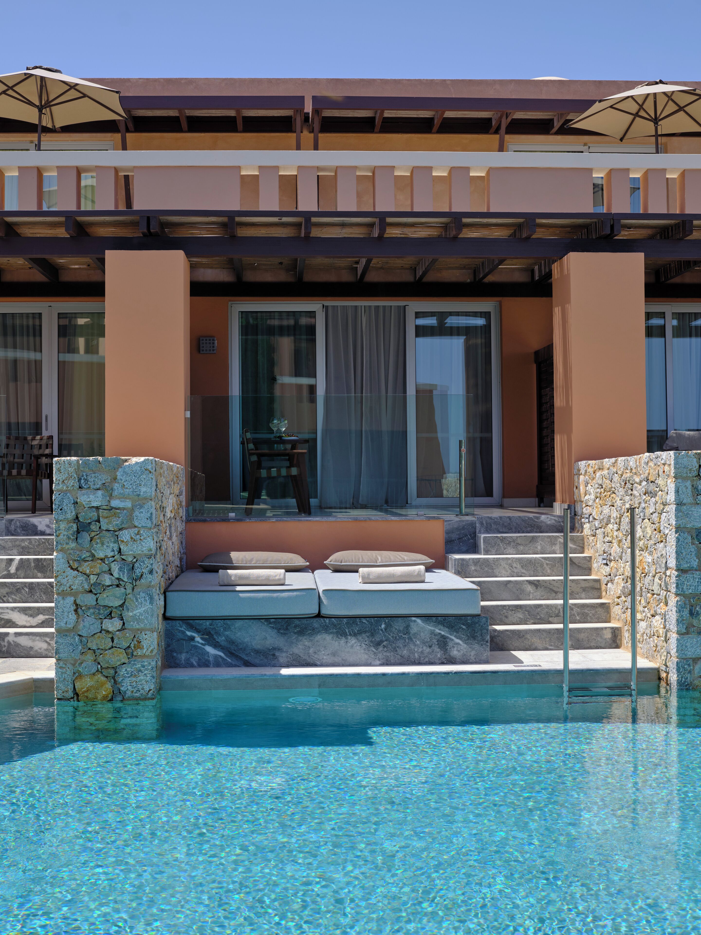 A pool area with a stone wall, a seating area with cushions, and a building with large windows and a covered patio.