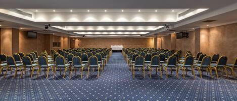 Un salón de conferencias con filas de sillas, un escenario y suelo alfombrado.