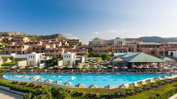 Un resort con piscina central rodeada de tumbonas y sombrillas, varios edificios hoteleros con un estilo arquitectónico uniforme, y montañas al fondo.