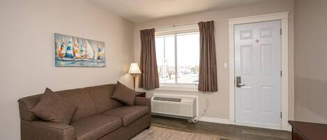 A living room with a brown sofa, a painting of sailboats, a window with curtains, a white door, and a ceiling light fixture.