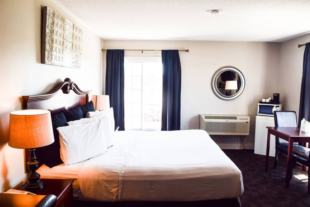 A hotel room with a bed, bedside lamp, air conditioning unit, desk, chair, and a round mirror on the wall.