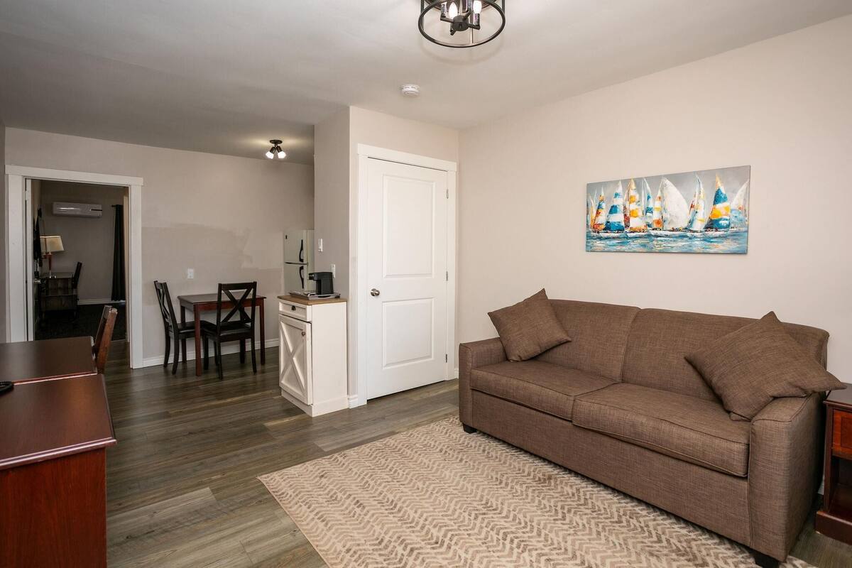 A living room with a brown sofa, a patterned rug, a painting of sailboats, and a dining area with a table and chairs.