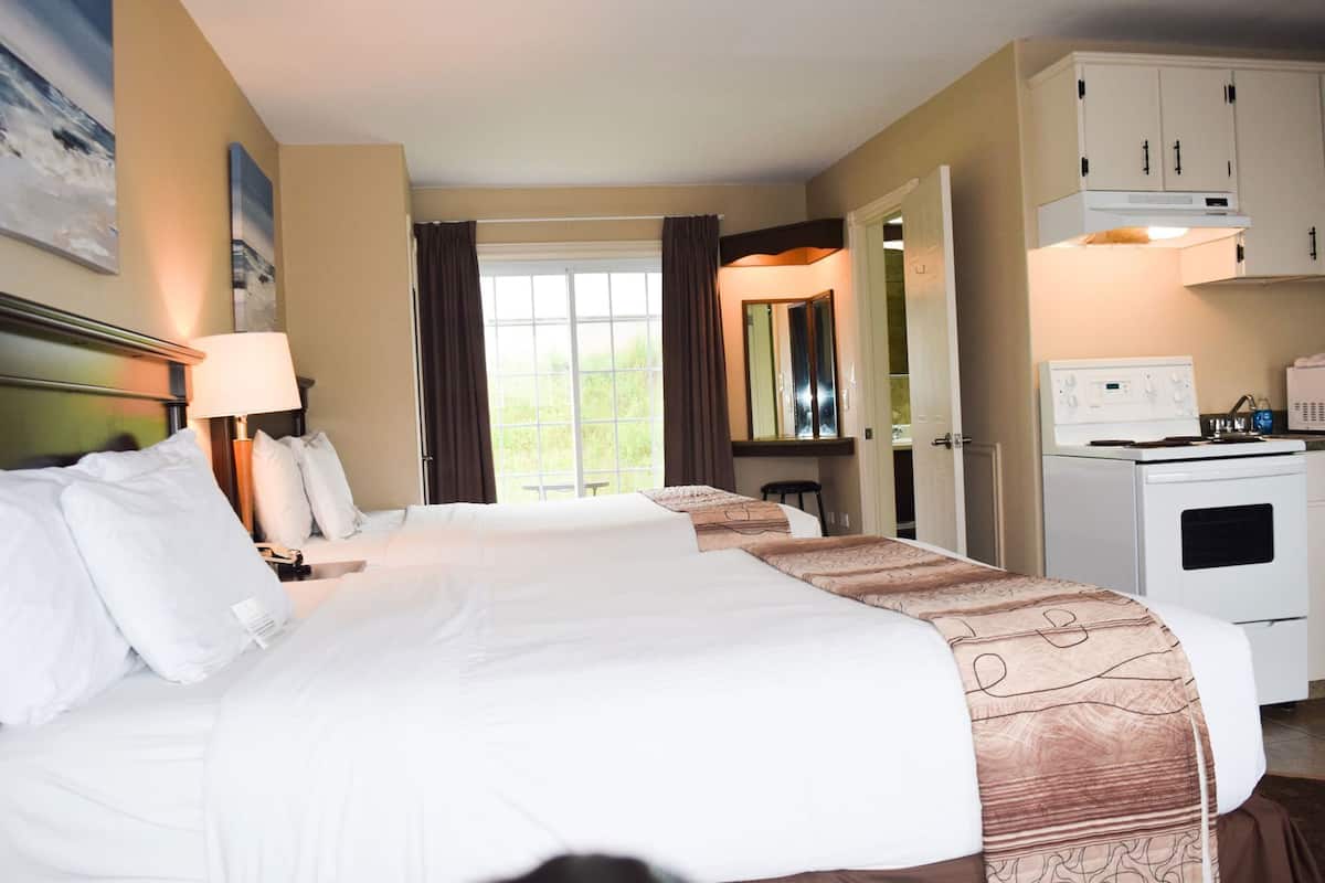 A hotel room with a large bed, bedside tables, a desk, a chair, a window with curtains, a kitchenette with a stove and microwave, and cabinets.