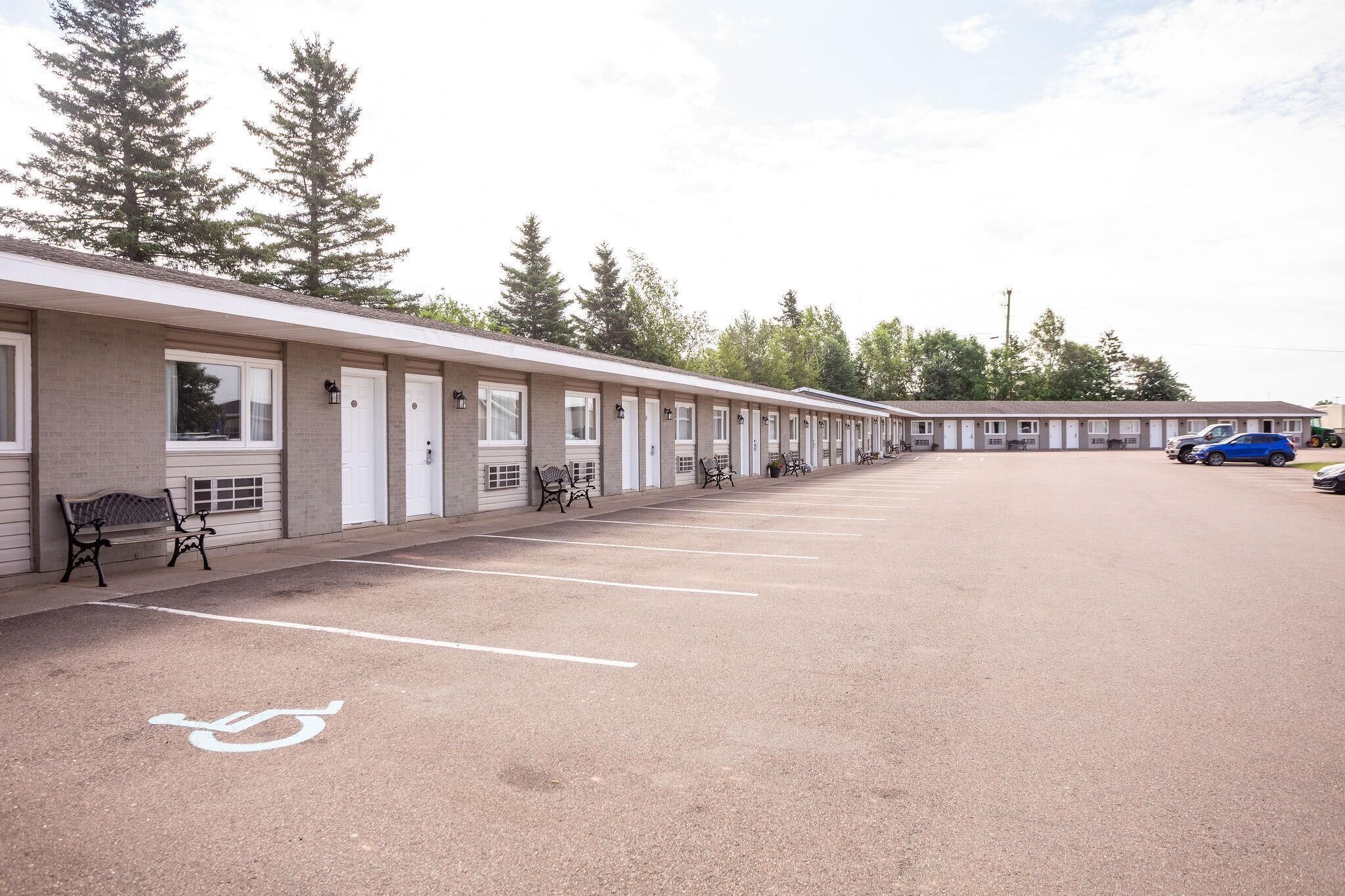 A motel with a row of single-story buildings, each with a front door and windows. There are parking spaces with a handicapped parking symbol, and a few benches along the walkway.