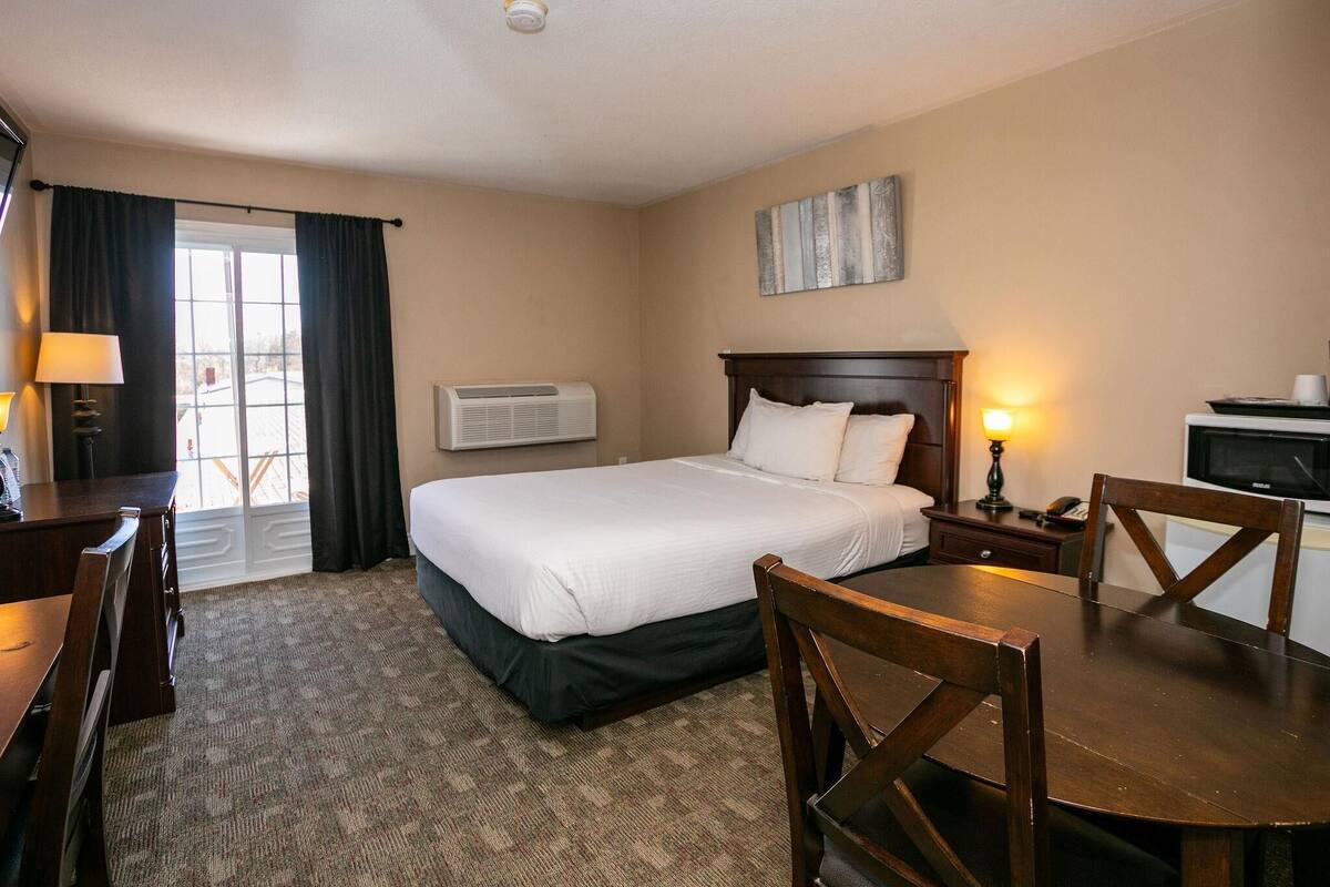 A hotel room with a bed, a desk, a chair, a table, a lamp, a TV, and a window with curtains.