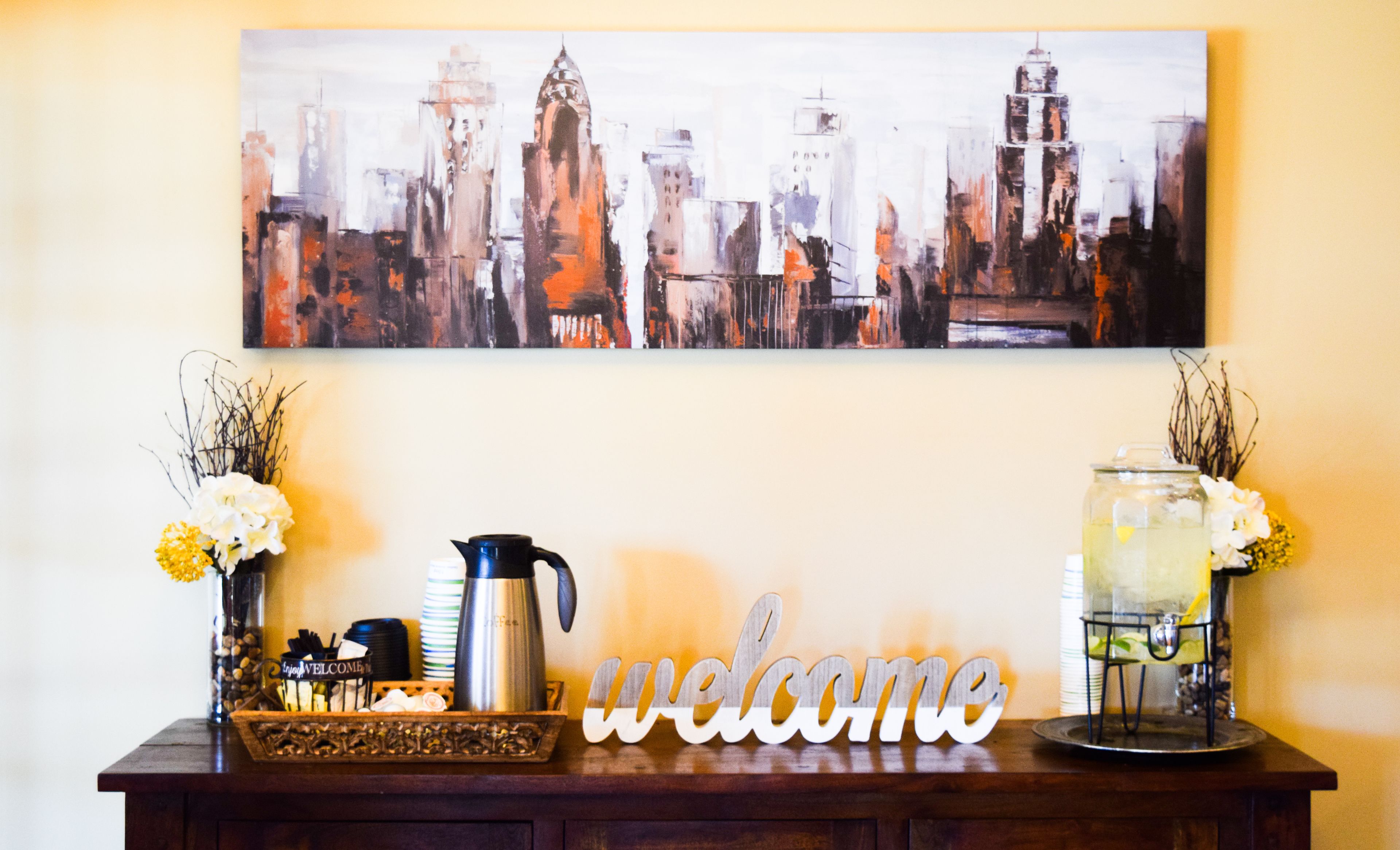 Een receptiegedeelte met een stadsgezicht schilderij, een bord met de welkomstwording, een koffiehoek en een decoratieve vaas met bloemen.