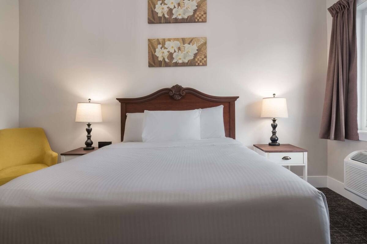 A hotel room with a large bed, two bedside tables with lamps, a yellow armchair, and artwork on the wall.