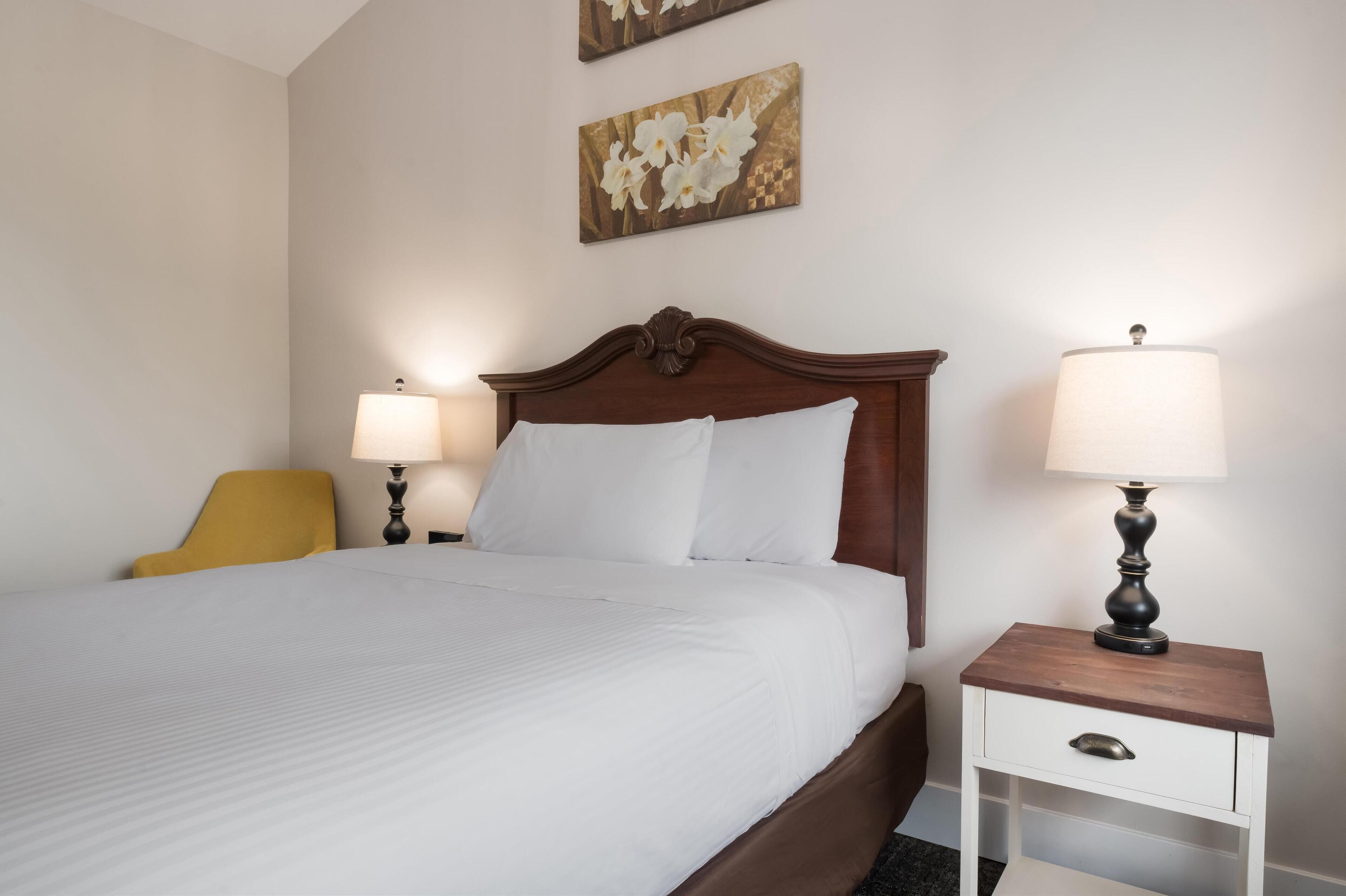 A neatly made bed with white linens, a wooden headboard, two bedside lamps, a small white nightstand with a drawer, and a mustard-colored chair.