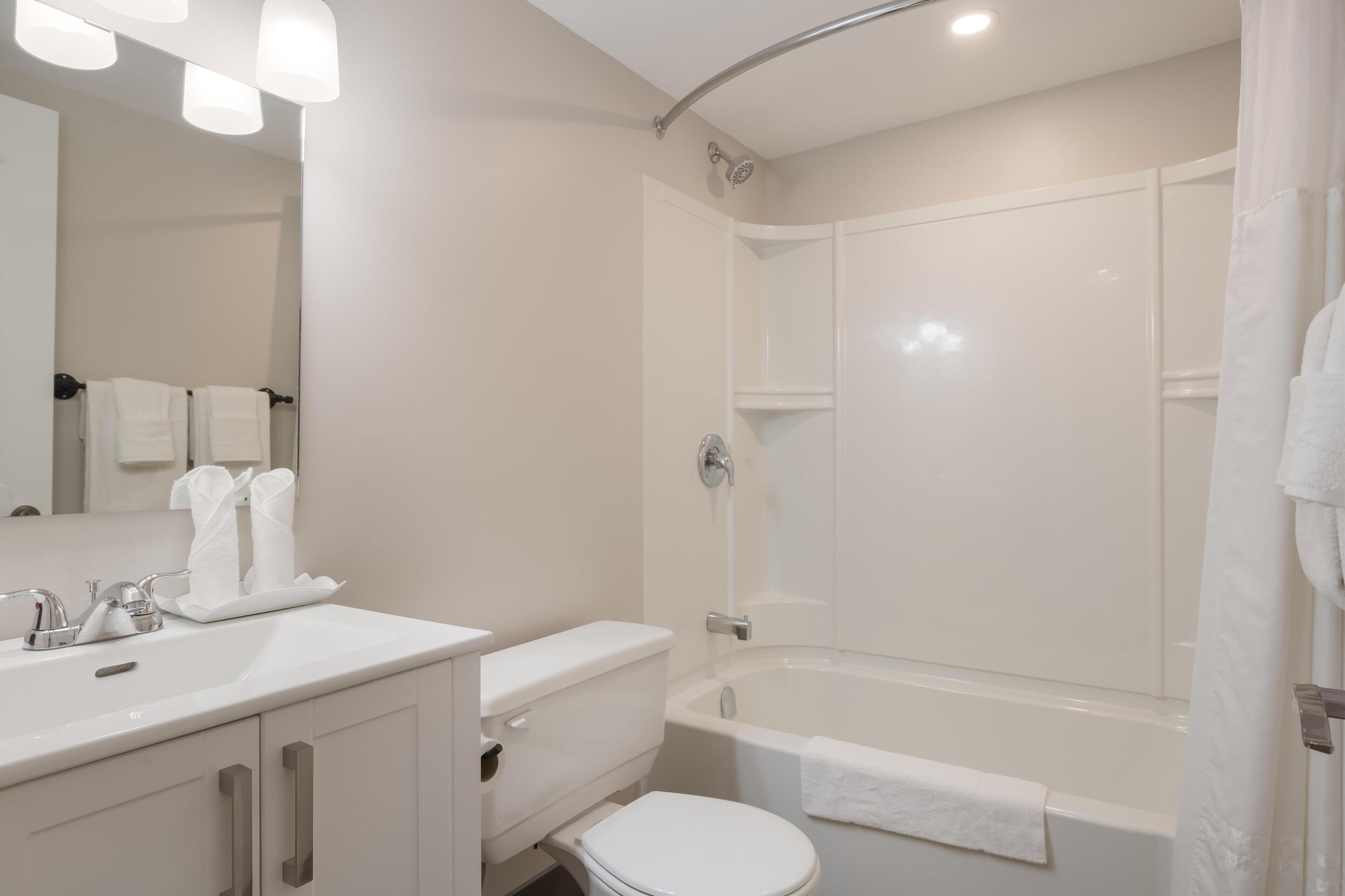 Une salle de bain moderne avec un lavabo, des toilettes et une baignoire, tous de couleur blanche. Des serviettes sont accrochées au mur et un rideau de douche est installé.