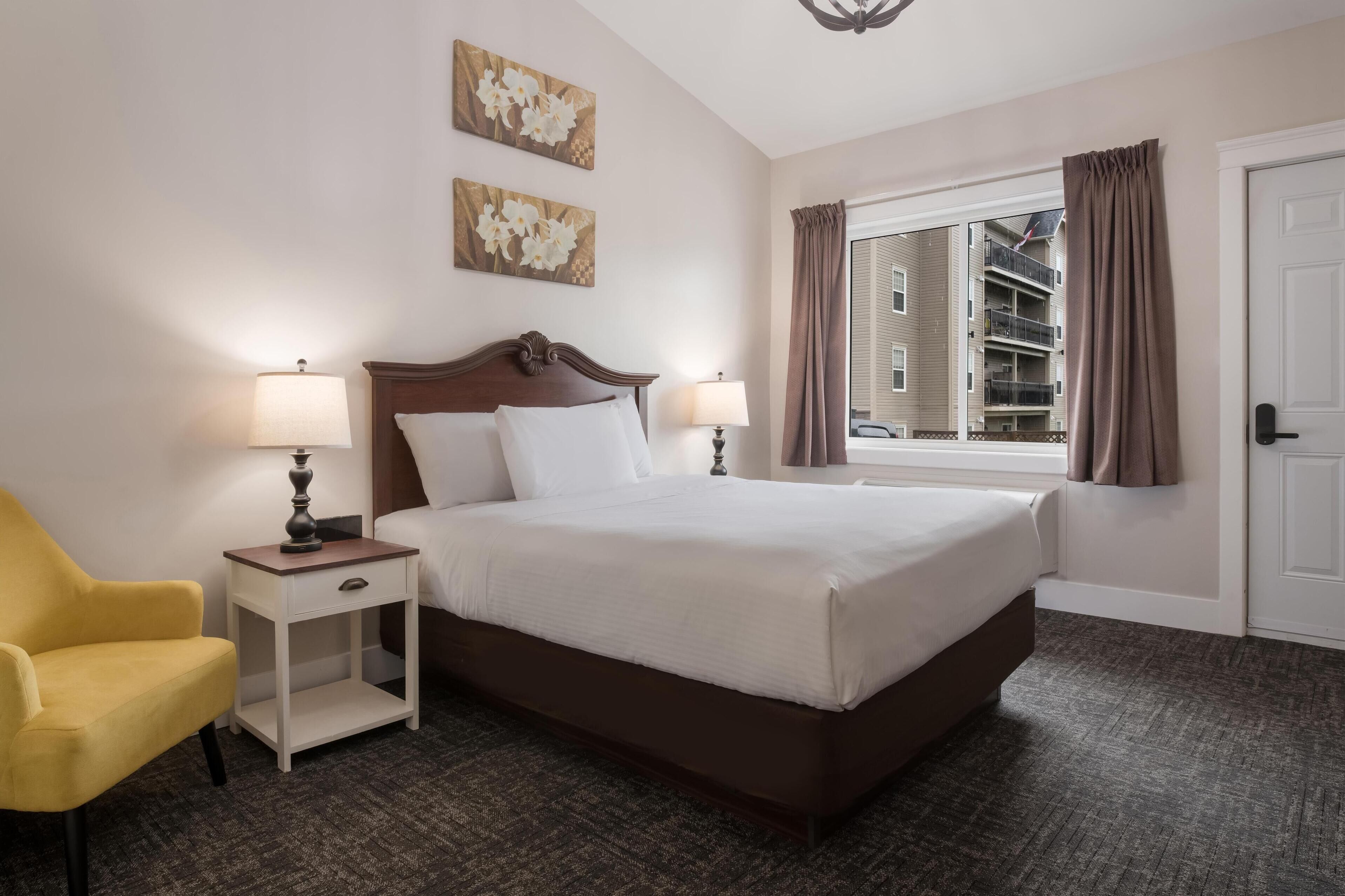Une chambre d’hôtel avec un grand lit, deux lampes de chevet, une table de chevet, un fauteuil jaune et une fenêtre avec des rideaux.