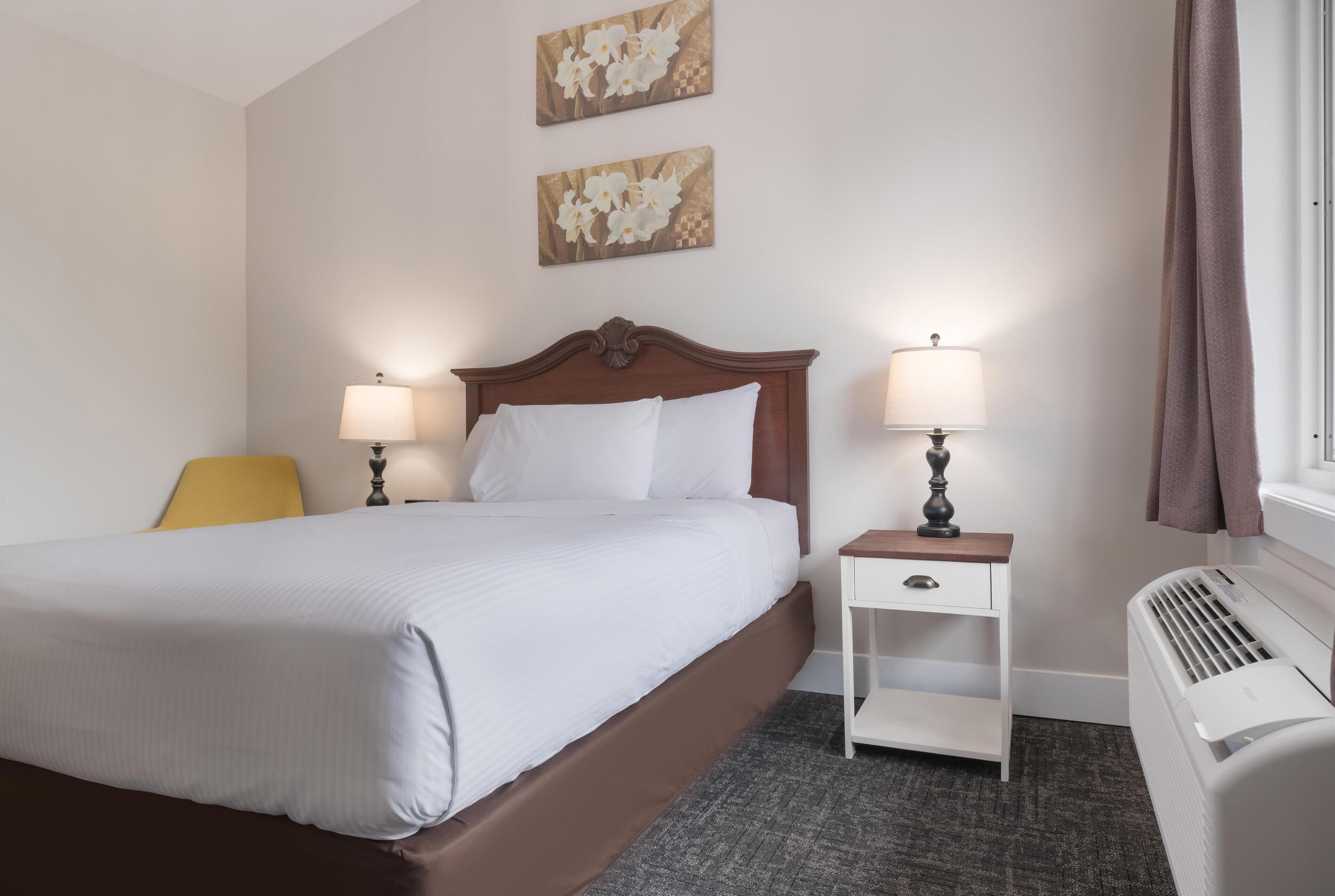 Ein Hotelzimmer mit einem großen Bett, zwei Nachttischen mit Lampen, einem kleinen weißen Beistelltisch, einem Heizkörper und zwei an der Wand hängenden Blumenbildern.