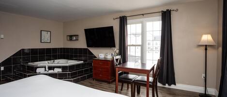 Habitación de hotel con una cama grande, un escritorio con silla, un aparador pequeño, un televisor de pantalla plana en la pared, una ventana con cortinas y una lámpara de pie.