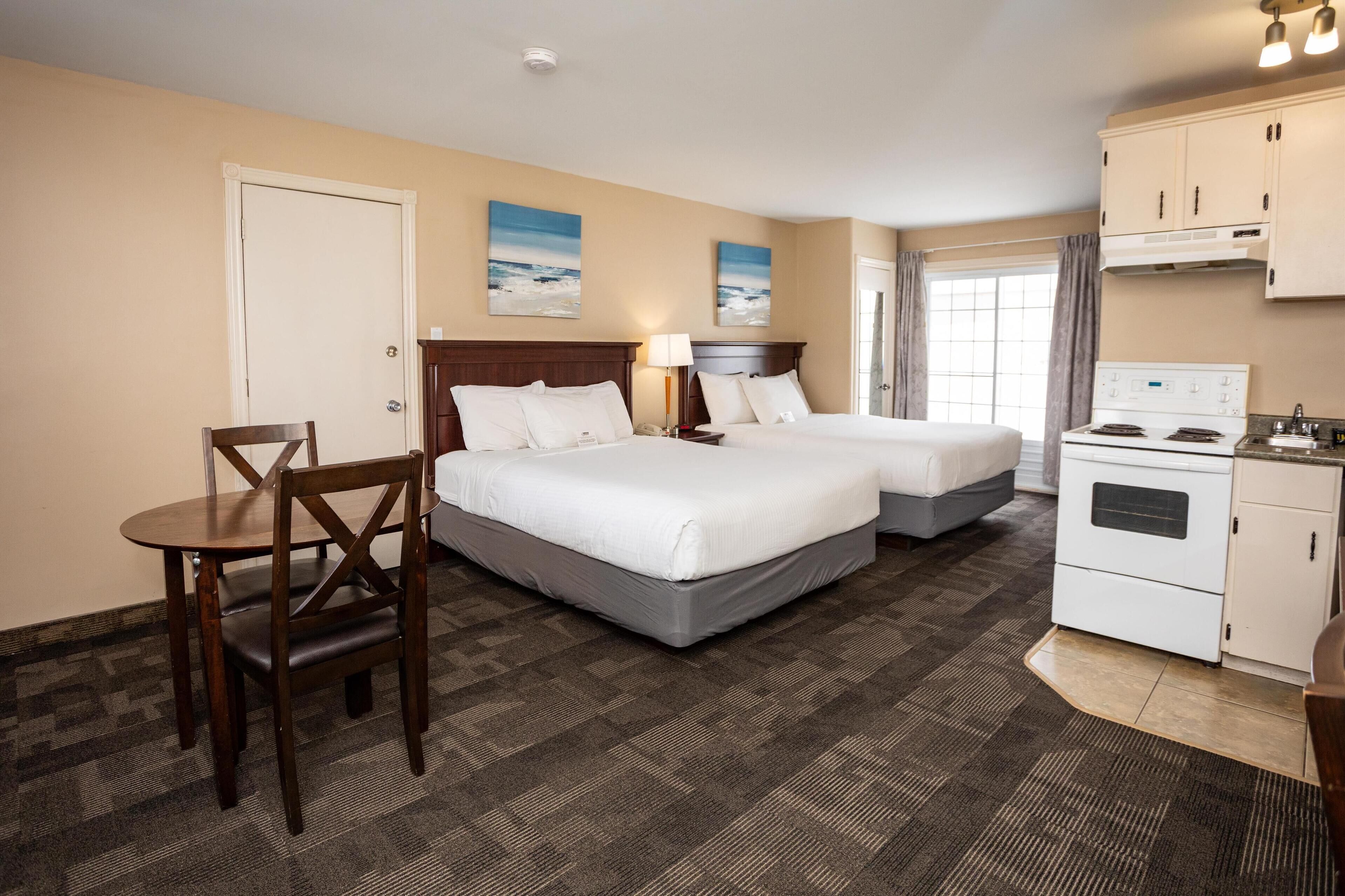 A hotel room with two beds, a desk, a chair, a kitchenette, and two paintings on the wall.