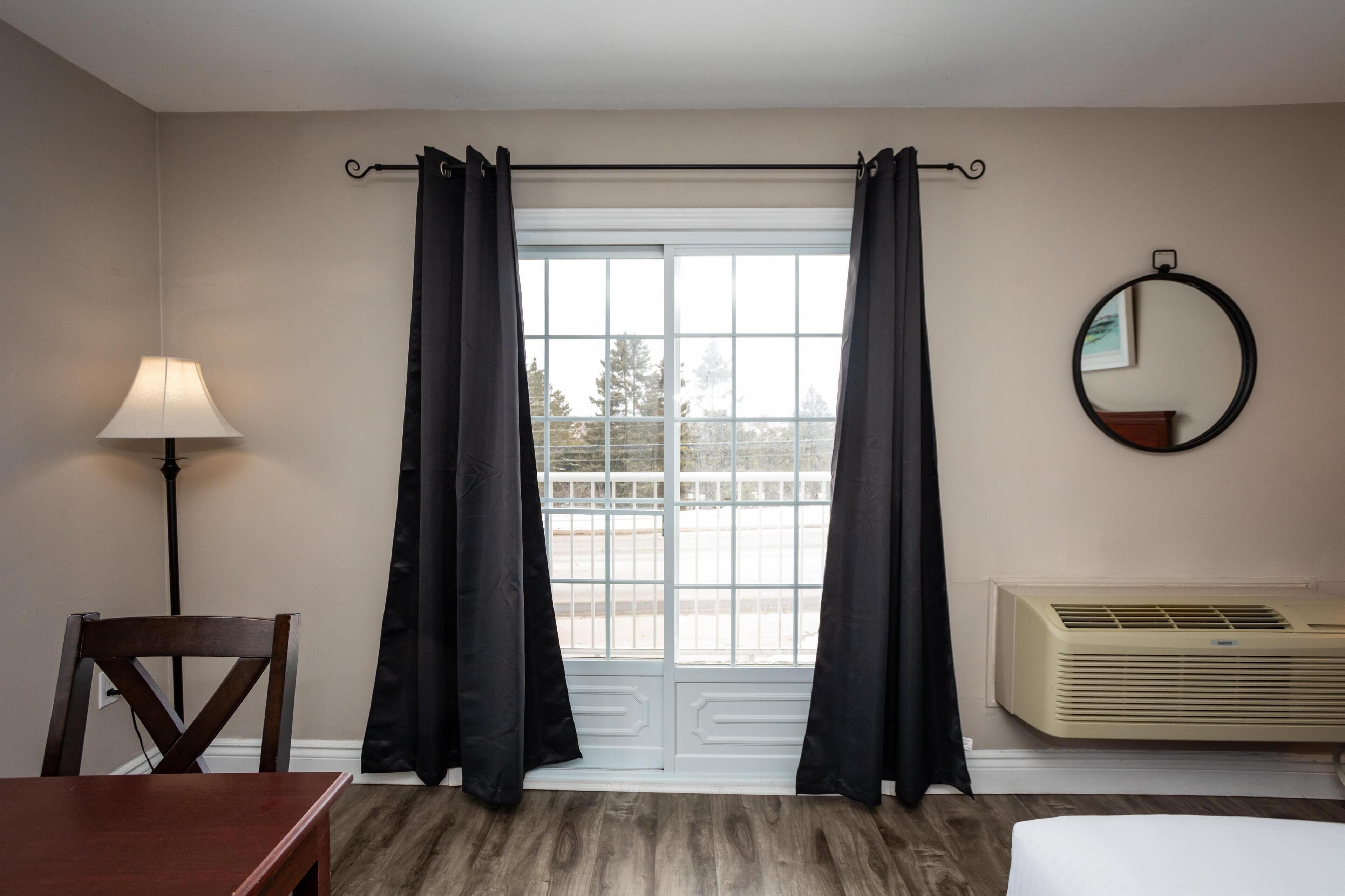 A hotel room with a wooden floor, a dining table, a chair, a lamp, a window with black curtains, an air conditioner, and a round mirror on the wall.