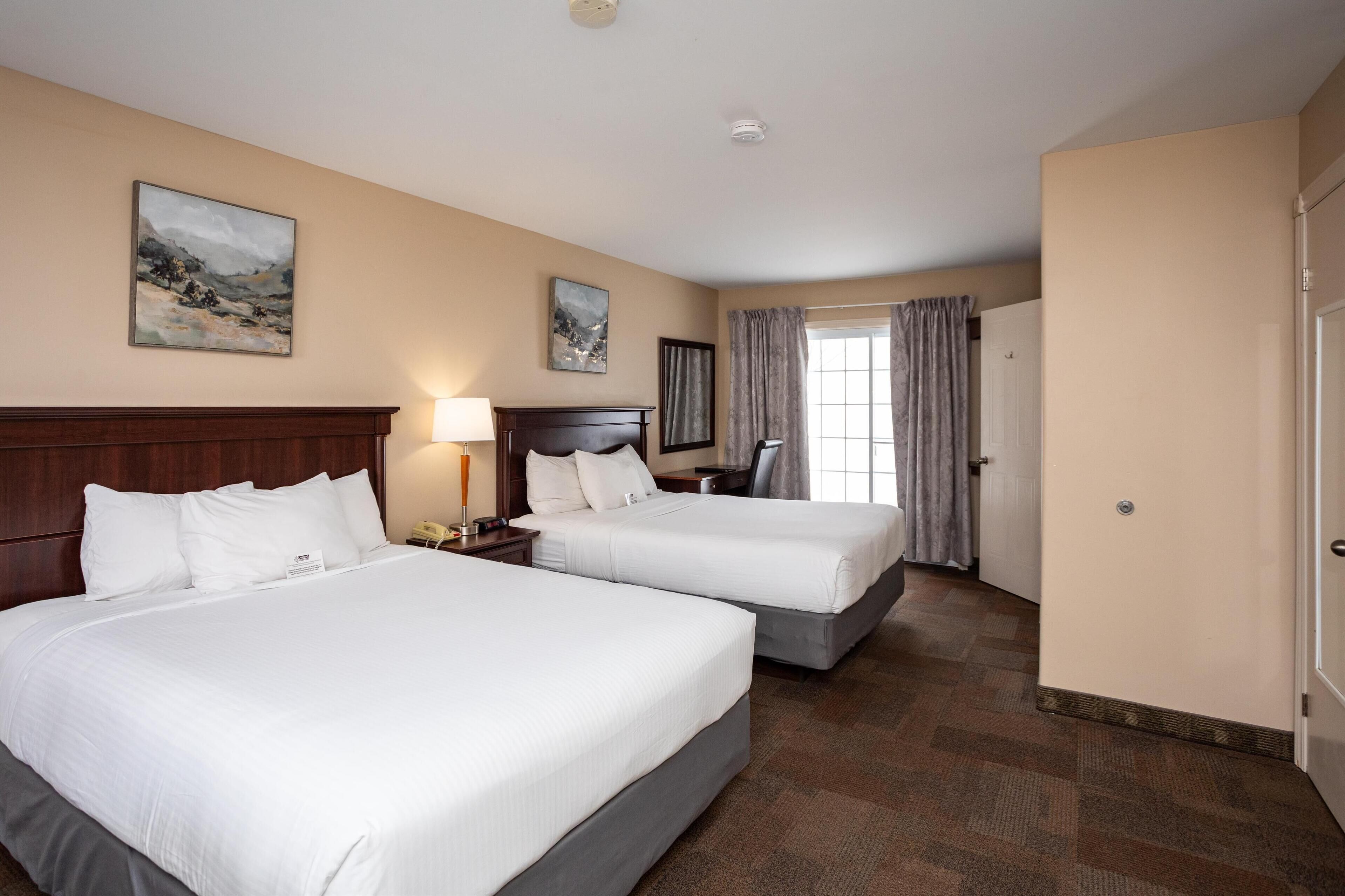 A hotel room with two beds, a desk, a chair, a lamp, and two paintings on the wall.