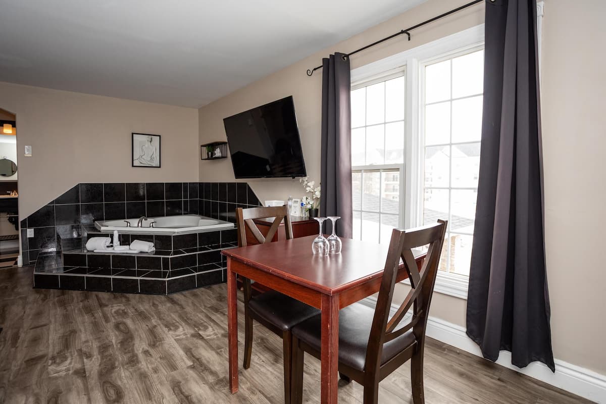 A modern kitchen with a built-in stove, a dining area with a wooden table and chairs, a large window with curtains, and a flat-screen TV mounted on the wall.