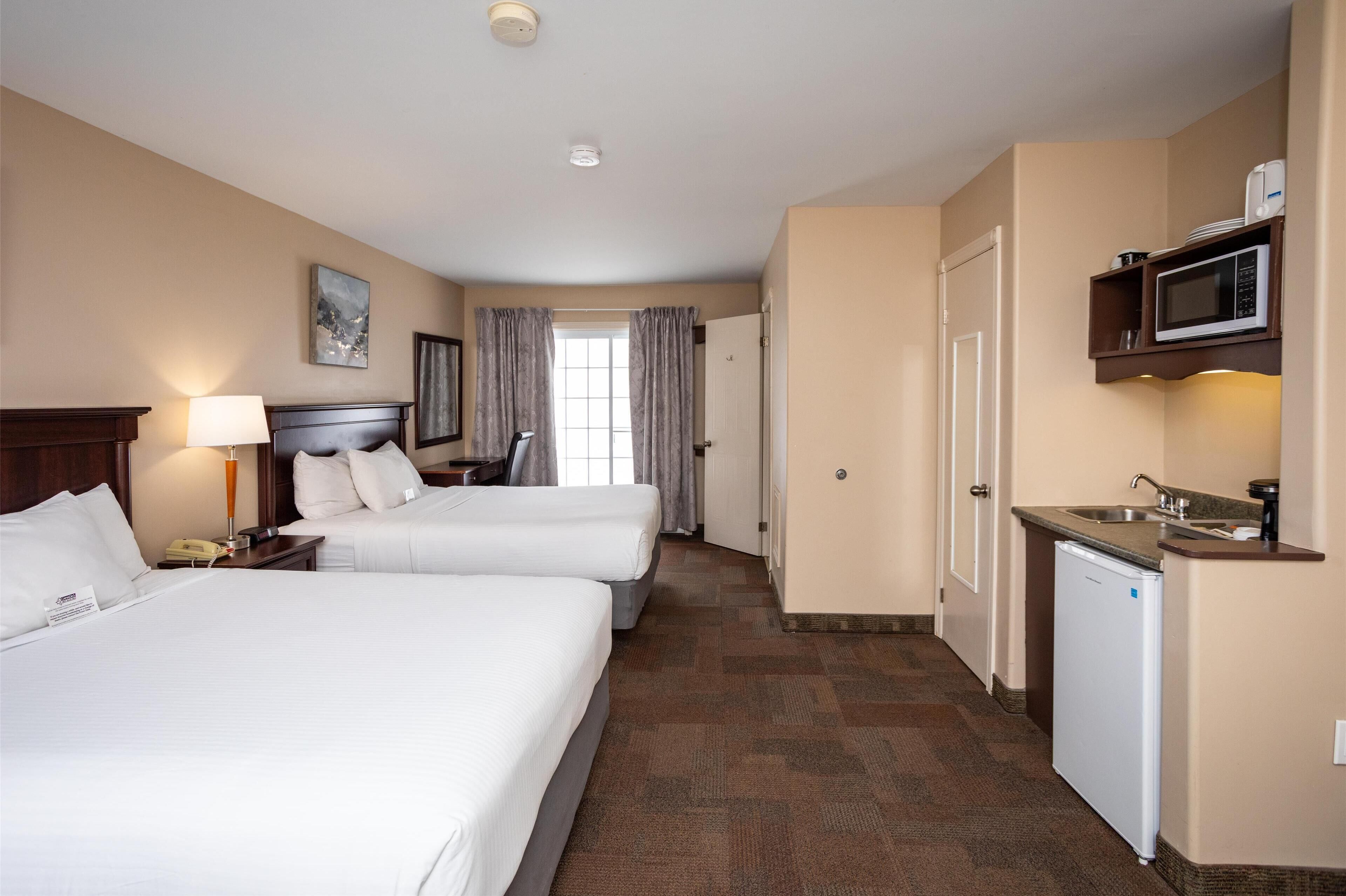 A hotel room with two beds, a desk, a lamp, a microwave, and a small kitchenette.